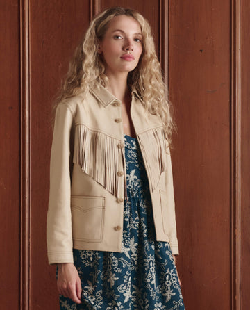 A woman with long curly hair wears THE GREAT. SP26 D1 "The Leather Fringe Jacket" in Toasted Almond over a blue and white floral dress, standing before a wooden panel wall.