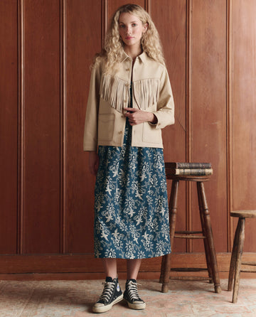 A woman stands indoors against wooden paneling, wearing THE GREAT. SP26 D1 The Leather Fringe Jacket in Toasted Almond, a blue patterned midi skirt, and black high-top sneakers. A stack of books sits on a small table beside her.