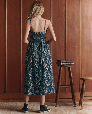 A woman with long blonde hair stands facing a wood-paneled wall, wearing THE GREAT. SP26 D1 The Verse Dress in Indigo Bandana and black sneakers. Two stacks of books and two wooden stools are nearby.