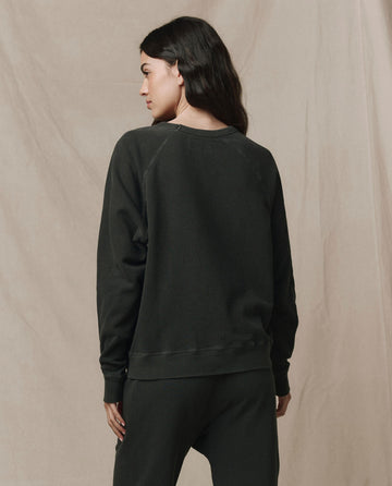 A woman stands facing away in THE GREAT. CORE KNITS The College Sweatshirt, Solid -- Washed Black, made from 100% cotton, with a beige fabric backdrop.