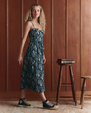 A woman wears THE GREAT.'s The Verse Dress in Indigo Bandana, styled with black high-top sneakers, standing indoors next to a wooden stool and stack of books against a wood-paneled wall.