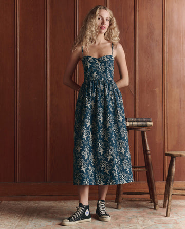 A woman stands indoors before wood paneling, wearing THE GREAT. SP26 D1’s The Verse Dress in Indigo Bandana, sculpted cotton with blue floral texture, paired with black high-top sneakers and resting one hand on books atop a stool.