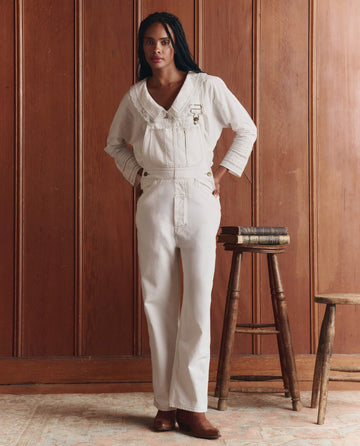 A person stands indoors by a wood-paneled wall, wearing THE GREAT. SP26 D1 The Union Slim Overall in Washed White with brown shoes, hands in pockets next to a wooden stool and stacked books.