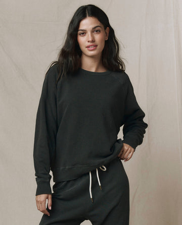 A woman poses against a neutral backdrop in THE GREAT. CORE KNITS' The College Sweatshirt in Solid Washed Black, pairing it with matching sweatpants and resting one hand on her hip.