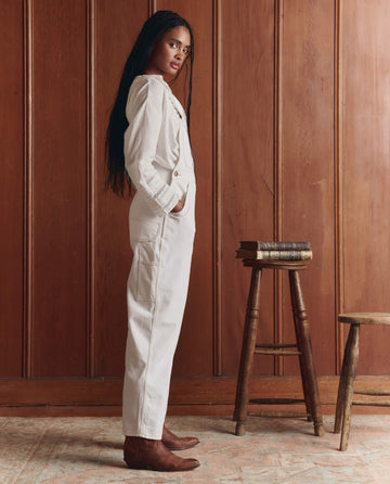 A woman in profile wears THE GREAT. SP26 D1 The Union Slim Overall in Washed White and brown boots, standing by a tall wooden stool with books, set before a wood-paneled wall.