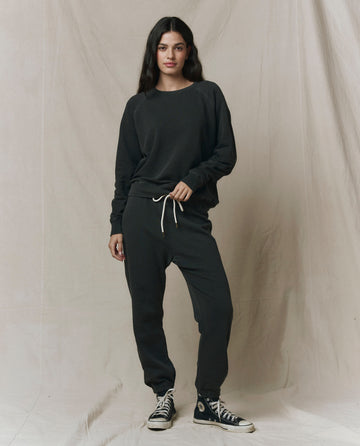 A woman stands against a neutral fabric backdrop, wearing THE GREAT. CORE KNITS The College Sweatshirt in Solid Washed Black—crafted from 100% cotton—and matching jogger pants with a drawstring, paired with black high-top sneakers.