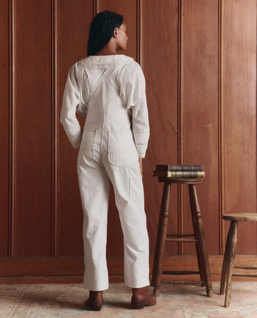 A person stands indoors facing a wooden wall, wearing THE GREAT. SP26 D1 The Union Slim Overall in Washed White, styled as vintage workwear with a long-sleeve shirt and brown boots. Books are stacked on a nearby wooden stool.