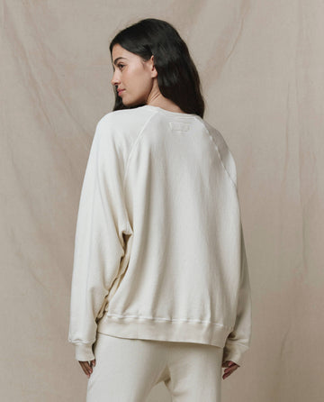 A woman with long dark hair models THE GREAT. CORE KNITS The Slouch Sweatshirt in Solid -- Washed White, standing with her back to the camera against a beige background.