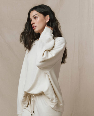 A woman wearing THE GREAT. CORE KNITS The Slouch Sweatshirt in Washed White stands against a neutral backdrop, touching her neck and glancing to the side.