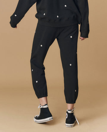 A person stands on a beige background wearing THE GREAT. PF25 EMB KNITS The Stadium Sweatpant. Embroidered in Almost Black With Daisy Embroidery, paired with a matching sweatshirt and black high-top sneakers.