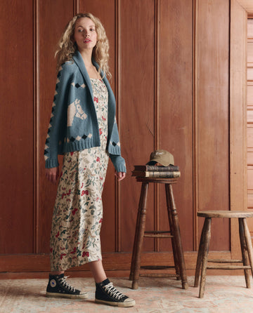 A woman in a floral dress, The Stable Lodge Cardigan in Blue Bell by THE GREAT. SP26 D1, and black sneakers stands beside a stool with stacked books and a cap against a wooden paneled wall.