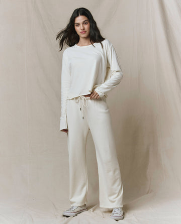 A woman stands against a neutral fabric backdrop, wearing THE GREAT. CORE KNITS The Long Sleeve Crop Tee in Washed White, paired with matching drawstring pants and white sneakers.