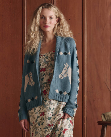 A woman with curly blonde hair wears THE GREAT. SP26 D1 Stable Lodge Cardigan in Blue Bell, featuring horse patterns, over a floral dress as she stands before a wooden paneled wall.