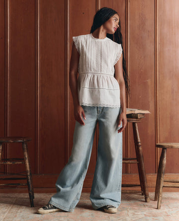 A woman stands indoors wearing THE GREAT. SP26 D1 The Sonnet Top in True White with lace pintuck details, wide-leg jeans, and black sneakers; wooden stools and a wood-paneled wall are in the background.