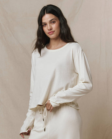 A woman with long dark hair wears THE GREAT. CORE KNITS' The Long Sleeve Crop Tee in Washed White, paired with matching pants, standing against a neutral background.