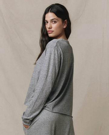 A woman with long dark hair wears THE GREAT. CORE KNITS' The Long Sleeve Crop Tee in Heather Grey, styled with matching pants, standing before a beige background and glancing over her shoulder.
