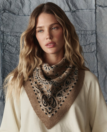 A woman with long wavy hair wears THE GREAT. H25 COLLECTION’s The Bandana Kerchief in Camel and a cream top, standing before a textured gray background.