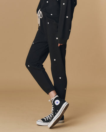 Someone wears THE GREAT. PF25 EMB KNITS The Stadium Sweatpant. Embroidered--Almost Black With Daisy Embroidery, paired with a matching black hoodie, white drawstring, and black Converse high-tops against a beige background.