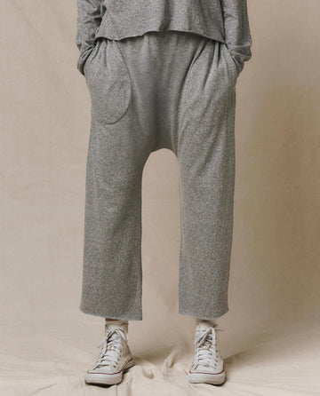 Wearing THE GREAT. CORE KNITS The Jersey Crop Pant in heather grey—loose-fitting straight-leg style with an elastic waist for comfort—paired with white high-top sneakers, hands in pockets, against a neutral background.