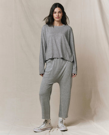 A person stands before a beige fabric backdrop wearing THE GREAT. CORE KNITS The Jersey Crop Pant in heather grey, paired with a matching loose-fitting long-sleeve top and white high-top sneakers.