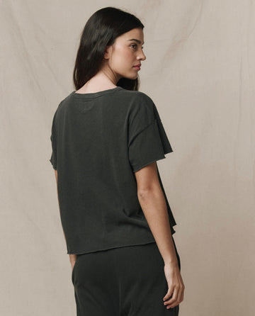 A woman with long dark hair stands sideways, facing left, wearing THE GREAT. CORE KNITS The Crop Tee in Solid Washed Black and matching pants against a neutral background.