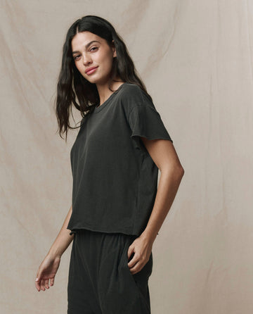 A woman with long dark hair wears THE GREAT. CORE KNITS The Crop Tee. Solid in Washed Black, paired with matching pants, standing before a neutral fabric backdrop.