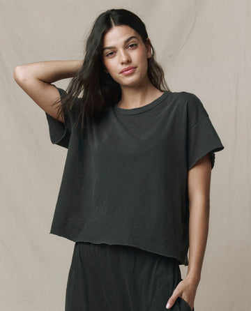 A woman with long dark hair wears THE GREAT. CORE KNITS The Crop Tee in Solid -- Washed Black, paired with matching pants, standing against a neutral background, one hand resting on her head.