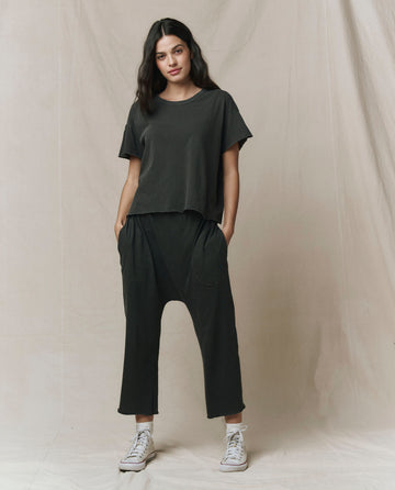 A woman stands against a neutral background wearing THE GREAT. CORE KNITS The Crop Tee. Solid -- Washed Black, styled with black drop-crotch pants and white sneakers, hands in her pockets. Super soft cotton. Made in USA.