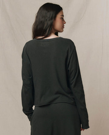 A woman with long dark hair stands facing away, wearing THE GREAT. CORE KNITS The Long Sleeve Crop Tee in Washed Black and a black skirt against a plain light background.