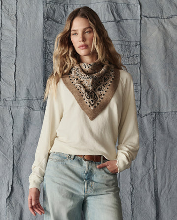 A woman in a cream sweater, light blue jeans, and THE GREAT. H25 COLLECTION's The Bandana Kerchief in Camel stands before a textured gray fabric backdrop.