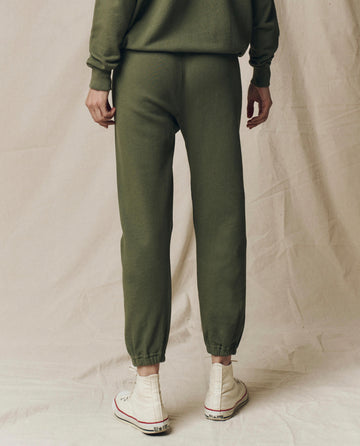 A person stands on a light fabric backdrop, wearing THE GREAT. SU25 SEASONAL KNITS' The Stadium Sweatpant. Solid in Heritage Army with a matching top and white high-top sneakers.