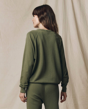 A person stands facing away in The College Sweatshirt. Solid -- Heritage Army by THE GREAT. SU25 SEASONAL KNITS, paired with matching pants, set against a beige fabric backdrop.