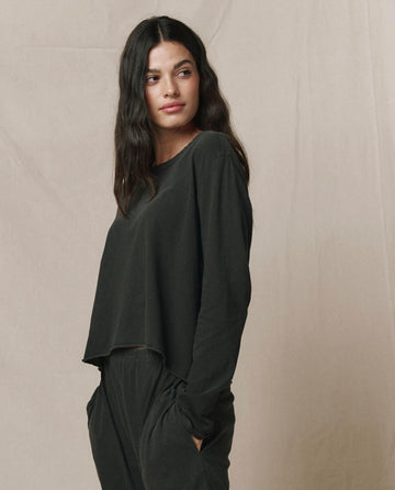 Woman with long dark hair wears THE GREAT. CORE KNITS The Long Sleeve Crop Tee in Washed Black, paired with matching black pants, standing with hands in pockets against a neutral background.