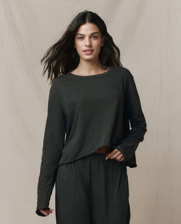 A woman with long dark hair wears THE GREAT. CORE KNITS The Long Sleeve Crop Tee in washed black, paired with matching pants, standing against a neutral background.