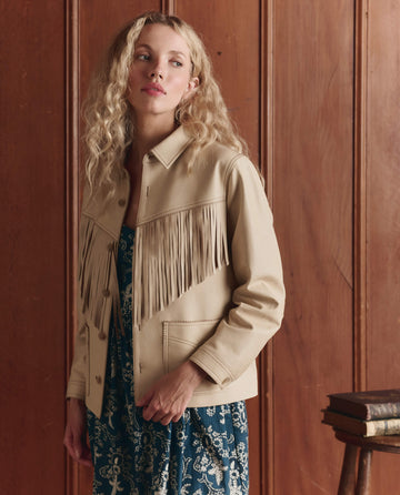 A person with long curly blonde hair wears THE GREAT. SP26 D1's The Leather Fringe Jacket in Toasted Almond over a blue patterned dress, standing indoors by a wooden panel wall and a small stack of books.