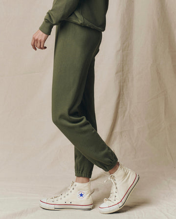 Model wears The Stadium Sweatpant. Solid in Heritage Army by THE GREAT. SU25 SEASONAL KNITS, paired with a matching sweatshirt and white high-top Converse, against a beige fabric backdrop.