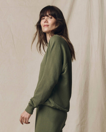 A person with long brown hair wears THE GREAT.'s The College Sweatshirt in Solid Heritage Army from the SU25 Seasonal Knits collection, paired with pants, standing against a beige fabric backdrop and facing left.