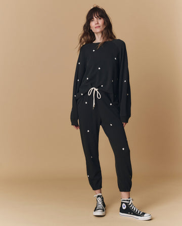 A woman stands against a beige background wearing THE GREAT. PF25 EMB KNITS The Stadium Sweatpant, Embroidered in Almost Black with Daisy Embroidery, paired with a black loose-fit sweatshirt and black high-top Converse sneakers.