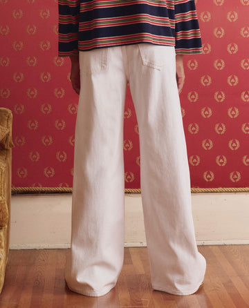 A person stands indoors, facing away, wearing THE GREAT. SP26 D1 The Slouch Wide Leg Jean in True White and a striped long-sleeve shirt. The scene includes red wallpaper with a gold pattern and a wooden floor.