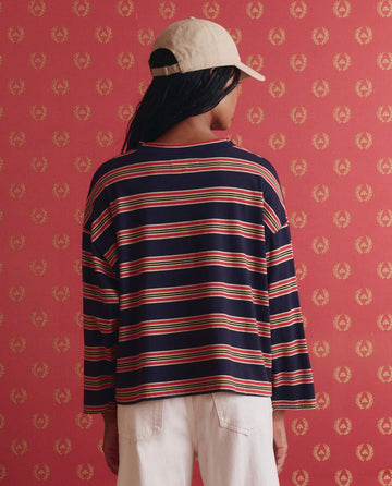 A person stands with their back to the camera, wearing THE GREAT. SP26 D1's The Field Tee in Scout Stripe, a beige cap, and white pants, set against red patterned wallpaper.