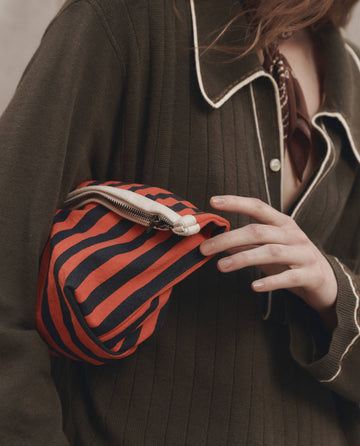 Wearing an olive green cardigan with a white-trimmed collar and a dark neck scarf, the person carries THE GREAT. F26 D1's The Stripe Pouch in Hot Red Mini Prep Stripe, tucked neatly under their arm.