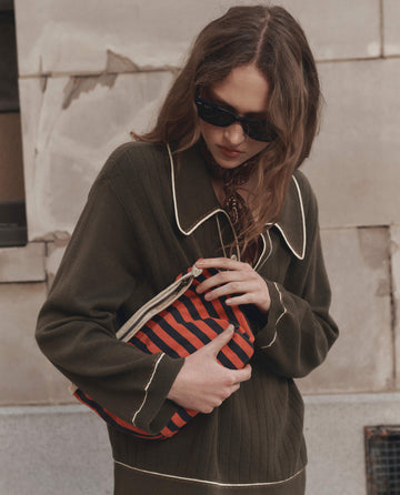 A woman with wavy hair wears black sunglasses, a patterned neckerchief, and an olive green knit shirt with contrast piping, holding THE GREAT. F26 D1's The Stripe Pouch in Hot Red Mini Prep Stripe in front of her.