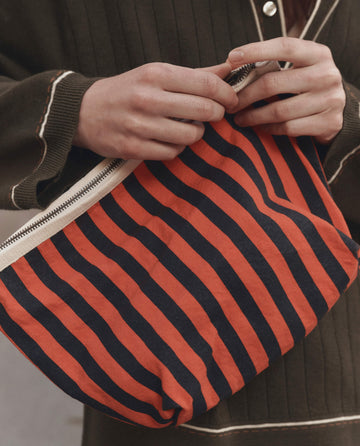 A person holding THE GREAT. F26 D1 The Stripe Pouch in Hot Red Mini Prep Stripe, styled with a dark long-sleeve top featuring white trim.