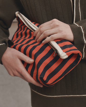 A person wearing a dark green sweater holds The Stripe Pouch in Hot Red Mini Prep Stripe by THE GREAT. F26 D1, featuring stylish diagonal red and navy stripes and a white zipper.