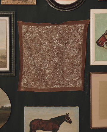 The Stetson Western Paisley Scarf in rust by THE GREAT. F26 STETSON—a brown square scarf featuring a white paisley pattern inspired by vintage bandanas—is displayed on a dark wall, surrounded by framed artwork and paintings, including images of horses.