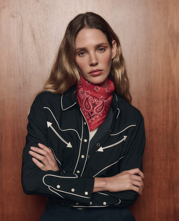 A woman with long hair stands against a wooden background, arms crossed, wearing THE GREAT. F26 COLLECTORS' The Western Shirt in black with white trim and a red paisley bandana.