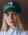A woman in a green THE GREAT. Baseball Hat with vintage panther embroidery and "The Great" text, paired with a light blue collared shirt, looks directly at the camera.