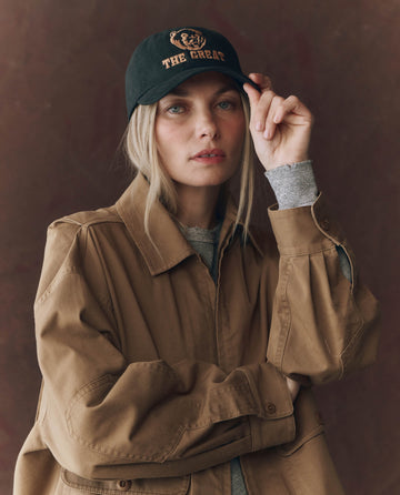 Wearing THE GREAT. F25 D1 ACCESSORIES The Baseball Hat, Graphic -- Black with chainstitch embroidery and an adjustable back, a person in a tan jacket looks at the camera while adjusting their cap.