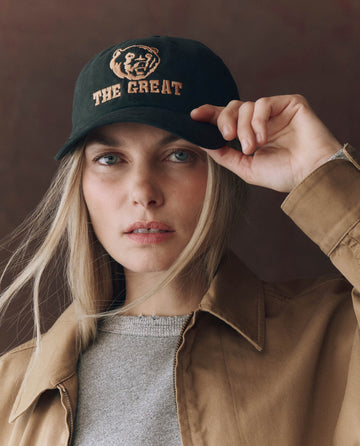 A person wears THE GREAT. F25 D1 ACCESSORIES' The Baseball Hat in Black, featuring a tiger logo and chainstitch "THE GREAT" embroidery, with a brown jacket and grey top, holding the cap's brim and looking at the camera.