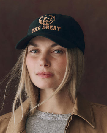 A woman with straight blonde hair wears THE GREAT. F25 D1 ACCESSORIES The Baseball Hat. Graphic -- Black, featuring "THE GREAT" chainstitch embroidery and an adjustable back, styled with a tan jacket and gray top against a plain background.